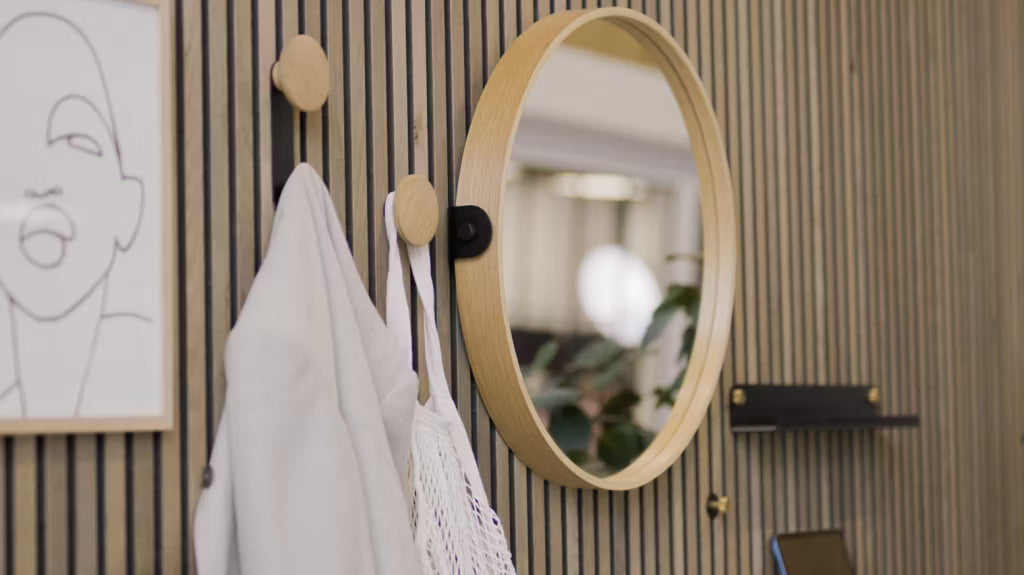 A round mirror mounted on a Nordic Akupanel wooden acoustic slat wall, white jackets hanging from heart-shaped hooks, and minimalist line art in the entryway.
